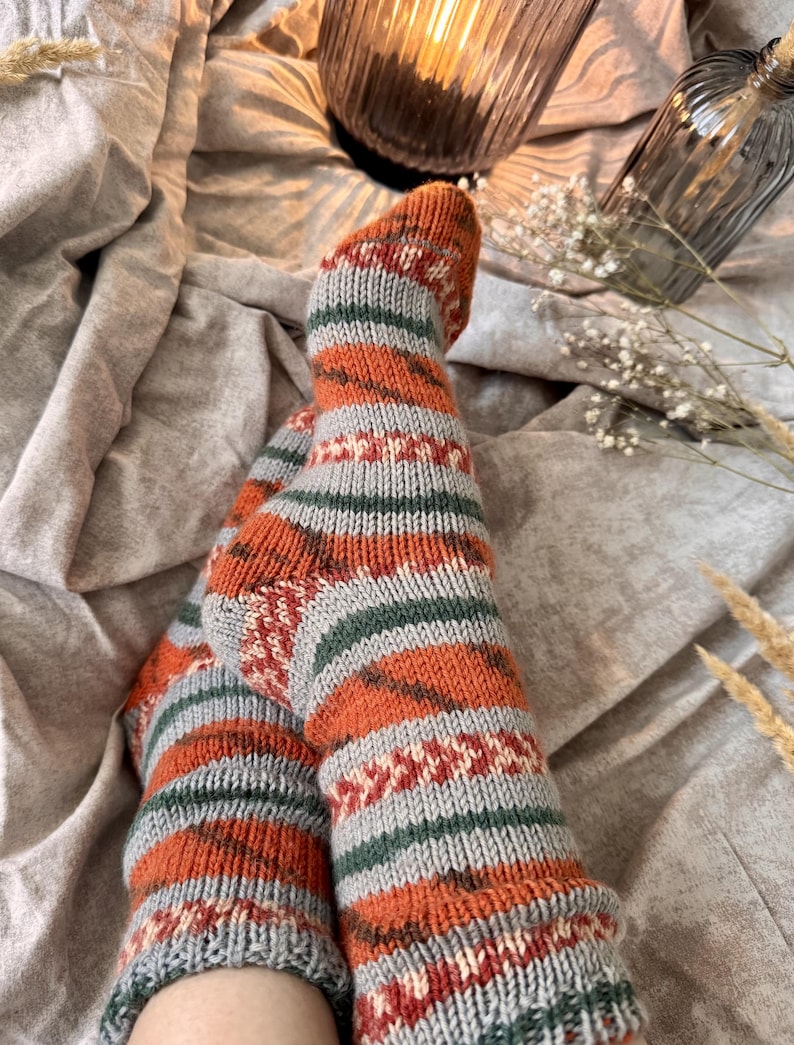 May include: A pair of hand-knitted socks featuring a horizontal striped pattern in burnt orange, light gray, and dark green. The design includes intricate geometric bands with small white accents, a ribbed cuff, and a reinforced heel construction.