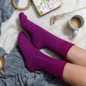 May include: A pair of purple socks with red stripes, worn by a person sitting on a white fluffy blanket. A cup of coffee and a book are also visible in the image.