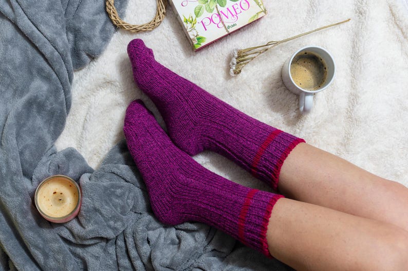 May include: A pair of purple socks with red stripes, worn by a person sitting on a white fluffy blanket. A cup of coffee and a book are also visible in the image.