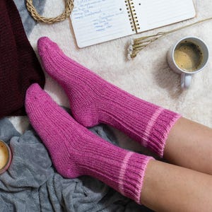 May include: A pair of pink knitted socks with white stripes, worn by a person relaxing on a white fuzzy blanket. A cup of coffee and a notebook are also visible.