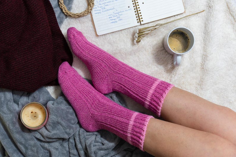 May include: A pair of pink knitted socks with white stripes, worn by a person relaxing on a white fuzzy blanket. A cup of coffee and a notebook are also visible.