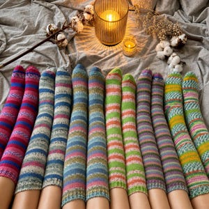 Op de afbeelding: Een collectie van twaalf gebreide sokken op een rij, elk met verschillende veelkleurige streeppatronen. De sokken tonen diverse kleurcombinaties waaronder roze, blauw, groen, geel en oranje, waarbij sommige sokken voorzien zijn van gestructureerde breipatronen.