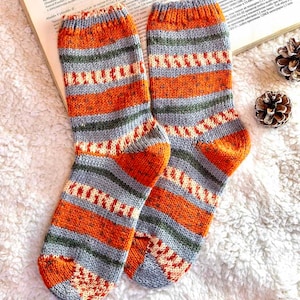 May include: A pair of hand-knitted wool socks featuring a horizontal striped pattern in shades of burnt orange, slate gray, olive green, and cream. The socks have a ribbed cuff at the top and a reinforced heel and toe section in solid orange.