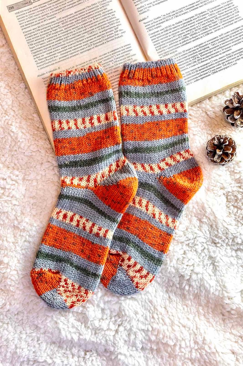 May include: A pair of hand-knitted wool socks featuring a horizontal striped pattern in shades of burnt orange, slate gray, olive green, and cream. The socks have a ribbed cuff at the top and a reinforced heel and toe section in solid orange.