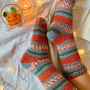 May include: A pair of hand-knitted socks featuring horizontal stripes in orange, grey, green, and cream. The knit pattern includes solid color blocks and textured, multi-colored sections. The socks have a ribbed cuff and a reinforced heel and toe.