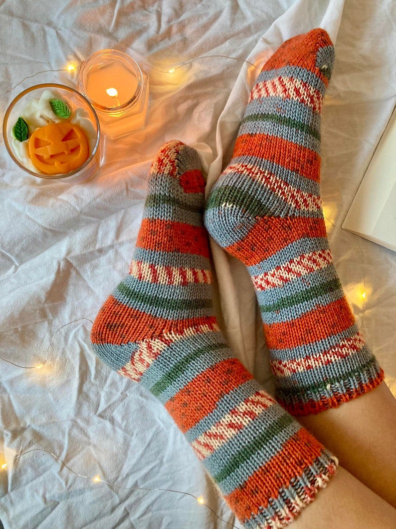 May include: A pair of hand-knitted socks featuring horizontal stripes in orange, grey, green, and cream. The knit pattern includes solid color blocks and textured, multi-colored sections. The socks have a ribbed cuff and a reinforced heel and toe.