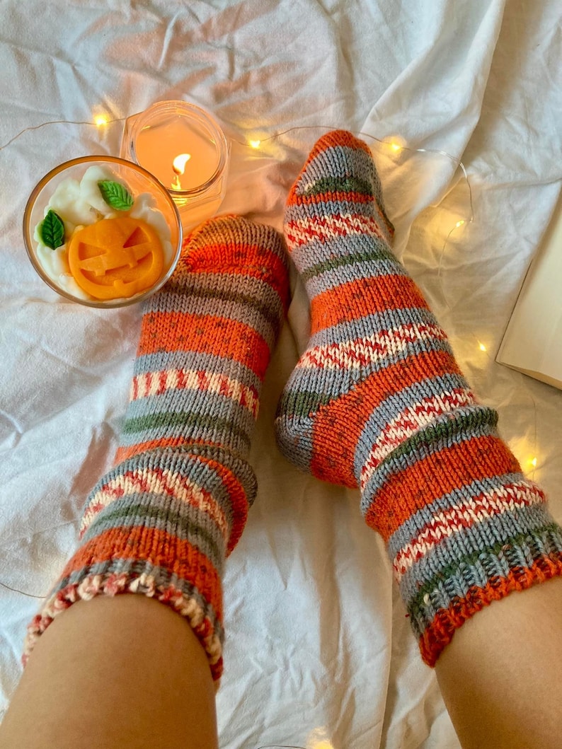 May include: A pair of hand-knitted socks featuring horizontal stripes in shades of orange, grey, white, and green. The socks have a textured knit pattern with speckled details throughout the yarn, extending from the ankle to the toe area.