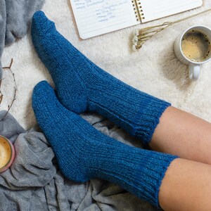 May include: A pair of blue knitted socks on a person's legs. The socks are ribbed and have a soft, cozy look. The person is sitting on a white fluffy blanket with a cup of coffee and a notebook nearby.