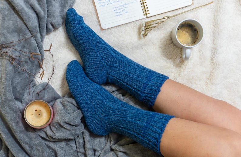 May include: A pair of blue knitted socks on a person's legs. The socks are ribbed and have a soft, cozy look. The person is sitting on a white fluffy blanket with a cup of coffee and a notebook nearby.