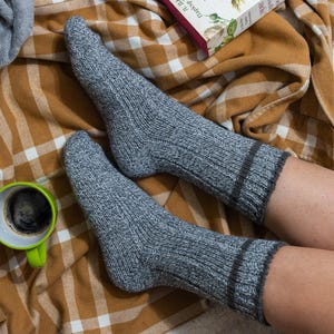 May include: A pair of gray knitted socks with a brown trim, worn by a person sitting on a plaid blanket with a green mug of coffee.