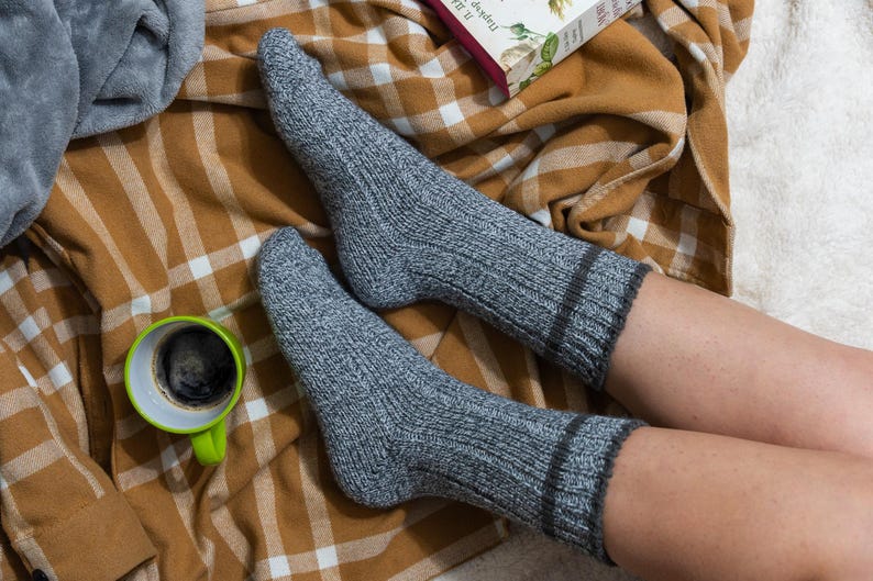 May include: A pair of gray knitted socks with a brown trim, worn by a person sitting on a plaid blanket with a green mug of coffee.