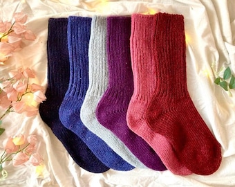 May include: Five pairs of knitted socks in different colours: purple, blue, grey, and red. The socks are folded and arranged in a row on a white fabric background.