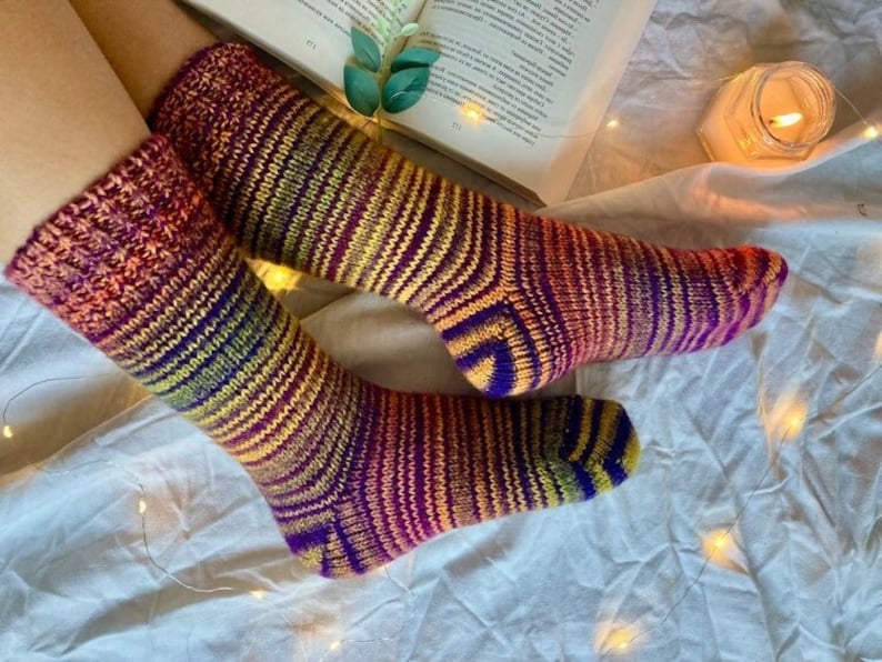 May include: A pair of colorful striped knitted socks in shades of purple, orange, yellow, and pink. The socks are worn on a person's feet and are resting on a white surface with string lights.