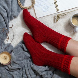 May include: A pair of red wool socks on a person's feet. The socks are on a white fluffy surface with a gray blanket and a cup of coffee.