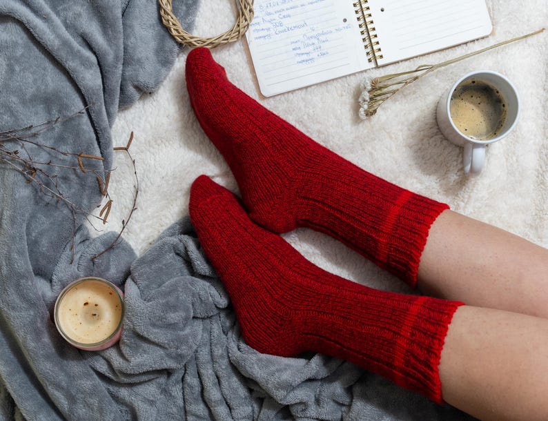 May include: A pair of red wool socks on a person's feet. The socks are on a white fluffy surface with a gray blanket and a cup of coffee.