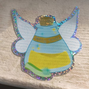 Puede incluir: Una botella de polvo de hada azul y amarilla con alas. La botella tiene un tapón de corcho y las palabras "Pixie Dust" escritas en ella.