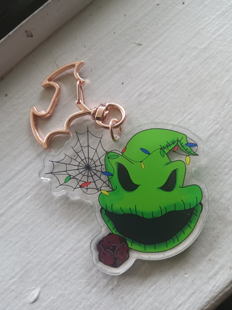 May include: A green and black character with a stitched mouth and a green hat with lights on it. The character is holding a red and black dice. The character is on a clear acrylic keychain with a gold metal bat-shaped charm.