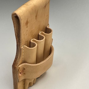 May include: A light tan leather tool holder with three cylindrical pockets and a curved back. The holder is secured with a copper rivet. The leather has a natural, unfinished appearance.