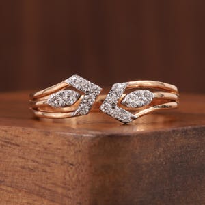 May include: Two rose gold rings with diamond accents. Each ring has a geometric design with a diamond-studded chevron shape and a central oval of diamonds. The rings are displayed on a wooden surface. The text "ROSYBLUEJEWELRY" is visible.