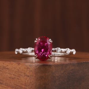 Könnte beinhalten: Ein silberner Ring mit einem großen, oval geschliffenen, tiefroten Edelstein, der von vier Krappen gehalten wird. Die Ringschiene ist mit mehreren kleinen, runden, klaren Steinen besetzt. Der Text ROSYBLUEJEWELRY ist auf der Innenseite sichtbar.