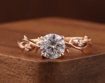 May include: A gold-colored ring with a large, round, clear gemstone at the center. The band features a vine-like design with small, clear accent stones. The ring is set on a wooden surface.
