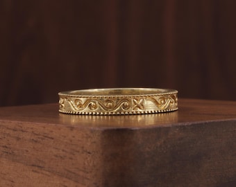 May include: A gold band ring with an intricate, repeating pattern of swirling lines and floral motifs. The ring is set on a dark wooden surface, showcasing its detailed design and warm color. The ring's surface has a textured appearance.