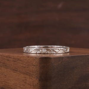 以下が含まれることがあります： 彫刻された花柄と3つの小さなダイヤモンドが付いたシルバーリング。リングは木製の表面に置かれ、その複雑なデザインと職人技が示されています。リングのバンドは、繊細で繰り返される葉とつるのモチーフが特徴です。