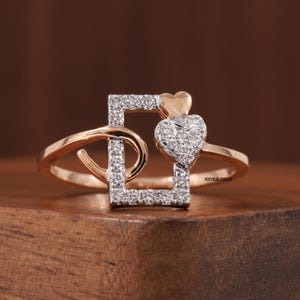 Puede incluir: Un anillo de oro rosa con un diseño cuadrado y de corazón. El cuadrado está incrustado con pequeñas piedras brillantes, y una forma de corazón está adornada con piedras similares. Un corazón más pequeño y de oro macizo se encuentra encima del corazón más grande.