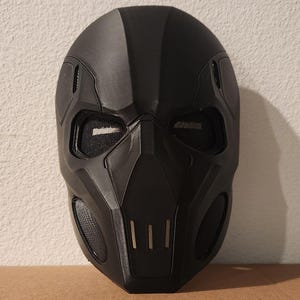 May include: A matte black full-face mask with a skull-like design. The mask features eye openings with mesh inserts, a segmented face, and vertical slits near the mouth. The overall design is angular and futuristic.