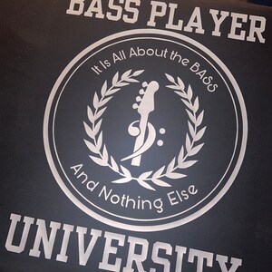 May include: Black t-shirt with white text that reads "Bass Player University". A white circle with a bass guitar and laurel wreath design reads "It is All About the Bass And Nothing Else".