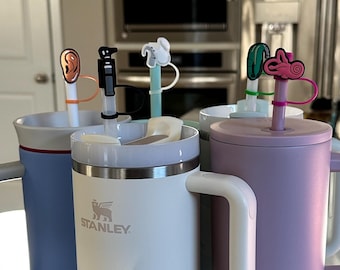 Puede incluir: Una colección de tazas de viaje aisladas en varios colores, incluyendo azul, blanco y lavanda. Cada taza tiene un asa y una tapa con una pajita. Las pajitas están rematadas con adornos decorativos, como una oreja, un robot y un conejo.