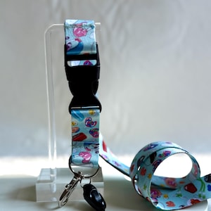 May include: A light blue lanyard with a repeating pattern of colorful cartoon images. The lanyard has a black plastic clasp and a metal clip. A black cord with a loop is attached to the lanyard.