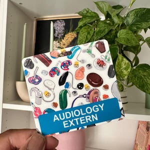 May include: A white card with a blue bar at the bottom that says "AUDIOLOGY EXTERN". The card is covered in colorful illustrations of hearing aids and other ear-related medical devices.