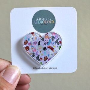May include: A heart-shaped acrylic charm with a white background and a colorful pattern of hearing aids and other audiology-related items. The charm is on a white card with the text "All Ears Audiology Etsy Shop" and "AllEarsAudiology.etsy.com".