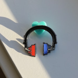 May include: A black headphone pin with red and blue earcups. The pin is set against a white background with a light blue heart-shaped object. The headphones are a stylized design.