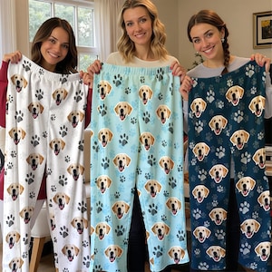 May include: Three pairs of pajama pants with a dog theme. The pants are white, light blue, and dark blue, each featuring a pattern of dog faces and paw prints. The pajama pants are made of a soft material.
