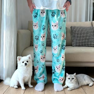 May include: Light blue pajama pants with a repeating pattern of cat faces and paw prints. The pants are worn by a person standing in front of a white dog and a gray cat.
