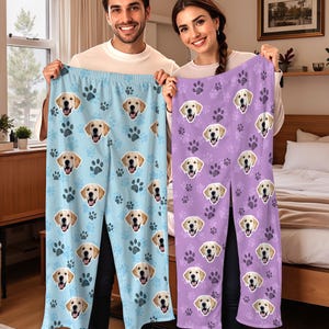 May include: Light blue and purple pajama pants featuring a repeating pattern of golden retriever faces and paw prints. The pants are held up by two people. The pajama pants are made of a soft, comfortable material.