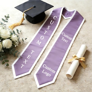 May include: A graduation cap with a gold tassel, two lavender custom text stoles with white trim, a rolled diploma tied with a gold ribbon, and a bouquet of white roses. The stoles read "Custom Text" and "Custom Logo".