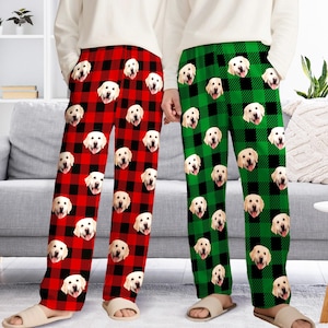 May include: Two pairs of pajama pants, one red and black plaid, the other green and black plaid. Both feature repeating images of a golden retriever's face. The pants are loose-fitting and have pockets.