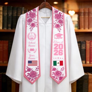May include: White graduation gown with a pink sash. The sash features floral designs, the text "School Name", "Your Name", the American flag, and "Class of 2026". The sash also has a graduation cap and diploma graphic.