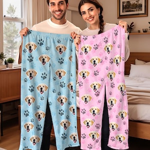 May include: Light blue and pink pajama pants featuring a repeating pattern of golden retriever faces and paw prints. The pants are held up by two people. The blue pants have dark blue paw prints, and the pink pants have dark pink paw prints.