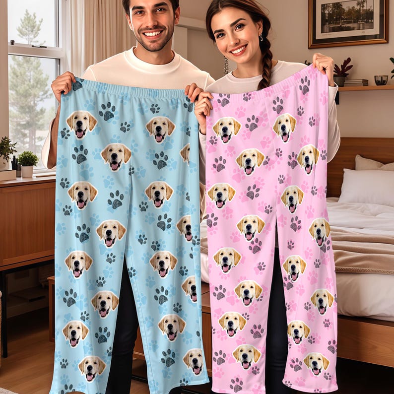 May include: Light blue and pink pajama pants featuring a repeating pattern of golden retriever faces and paw prints. The pants are held up by two people. The blue pants have dark blue paw prints, and the pink pants have dark pink paw prints.
