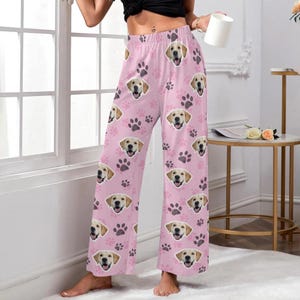 May include: Pink pajama pants featuring a repeating pattern of smiling golden retriever faces and paw prints. The wide-leg pants are made of a soft material and are suitable for lounging or sleeping. The pants are a light pink color.