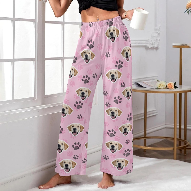 May include: Pink pajama pants featuring a repeating pattern of smiling golden retriever faces and paw prints. The wide-leg pants are made of a soft material and are suitable for lounging or sleeping. The pants are a light pink color.