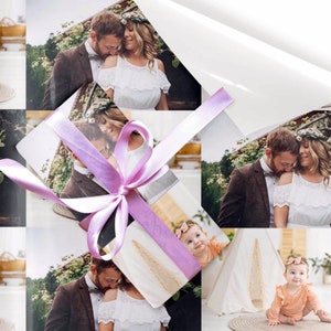 May include: A gift wrapped in white wrapping paper with a repeating pattern of photographs. The photographs show a couple and a baby. The gift is tied with a light purple ribbon.