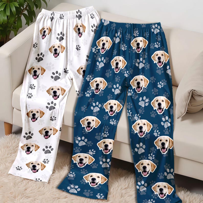 May include: Two pairs of pajama pants, one white and one navy blue, are displayed. Both feature a repeating pattern of golden retriever faces and paw prints. The pants are made of a soft material and are designed for comfort.