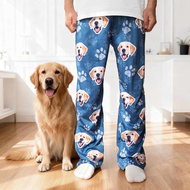 May include: Blue pajama pants with a repeating pattern of golden retriever faces and paw prints. A golden retriever dog sits next to the pants. The pants are worn by a person wearing white socks and a white shirt.