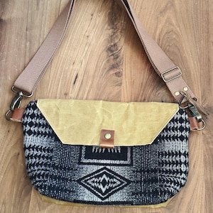 Puede incluir: Un bolso bandolera de lona marrón con una solapa con estampado geométrico en blanco y negro. El bolso tiene una correa de cuero marrón y una hebilla de latón.