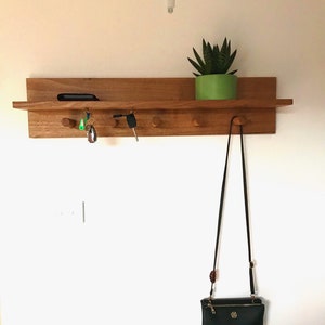 May include: A wooden wall-mounted shelf with hooks for hanging items. The shelf has a small plant in a green pot on top and keys and a handbag hanging from the hooks.